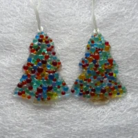 Two colourful beaded glass Christmas tree suncatchers, laying on a white cloth, hanging from white ribbons.
