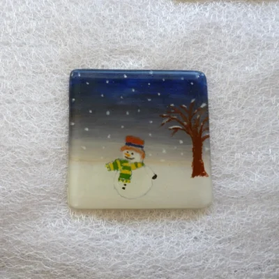Single square fused glass snowman coaster. Night sky with snow falling, snowman with red hat and green and yellow scarf. Tree on the right side.