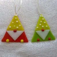 Two fused glass gonks, one red and yellow, the other green and yellow laying on a white cloth. Hangs from a white ribbon.