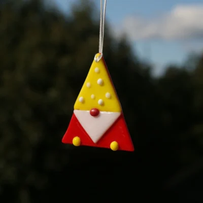 Triangle shaped fused glass gonk in yellow and red, white beard, red nose, yellow feet, white beads on yellow hat. Hanging from a white ribbon