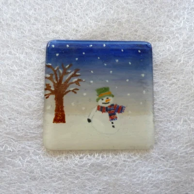Single square fused glass snowman coaster. Night sky with snow falling, snowman with green hat and blue and red scarf. Tree on the left side.