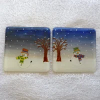 Fused glass snowmen coasters laying side by side on a white cloth. Blue sky, white snow with snowmen in the lower middle of the coaster.