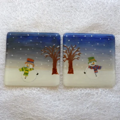 Fused glass snowmen coasters laying side by side on a white cloth. Blue sky, white snow with snowmen in the lower middle of the coaster.