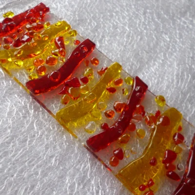 Close up of very colourful red yellow orange suncatcher showing tactile glass and glass frit laying on white cloth.
