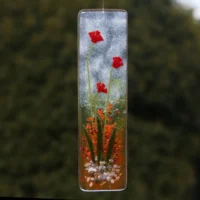 Long fused glass suncatcher with tactile red flowers and green stems and leaves. Background of suncatcher is white, yellow and red. Hanging from a clear cord.