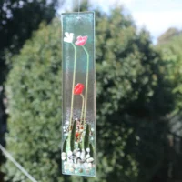 Long and slim fused glass flower suncatcher. Tactile red and white flowers, along with green glass leaves and stems fused onto a base glass. Hanging from a clear cord.