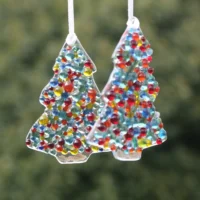 Two small colourful handmade fused glass Christmas tree suncatchers. Colourful glass beads green, yellow, orange, blue, turquoise fused on clear glass, hanging from white ribbons.