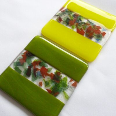 Two square unique handmade fused glass coasters in vibrant yellow and refreshing green, with centre decoration of clear glass and confetti glass. Bumpers on the back.