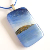Blue fused glass seascape pendant laying at an angle with sterling silver bail and blue cord necklace