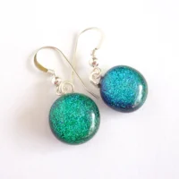 Handmade green dichroic glass earrings laying at an angle on a white background. Silver plated bails attached to green round cabochons and dangle from sterling silver hooks.