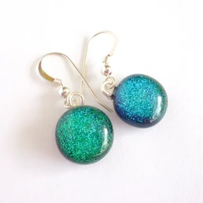 Handmade green dichroic glass earrings laying at an angle on a white background. Silver plated bails attached to green round cabochons and dangle from sterling silver hooks.