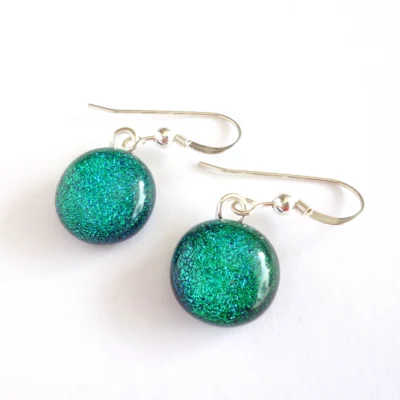 Green dichroic glass earrings laying at an angle. Round cabochons hanging from silver plated bails and 925 sterling silver ear hooks.