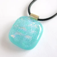 Turquoise blue dichroic glass pendant laying at a slight angle on a neutral background. 925 sterling silver bail and black cord necklace attached.