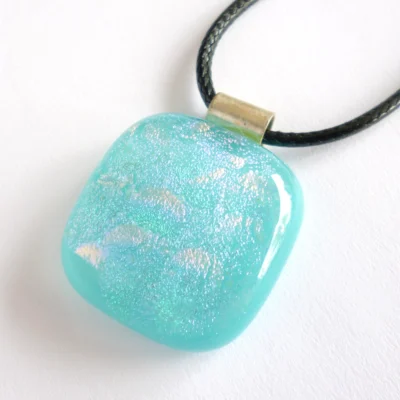 Turquoise blue dichroic glass pendant laying at a slight angle on a neutral background. 925 sterling silver bail and black cord necklace attached.
