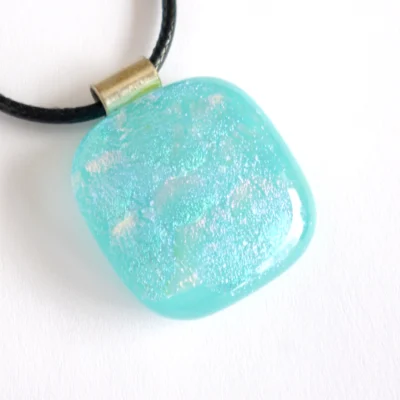 Close up and at an angle of turquoise blue dichroic glass pendant with shimmers of blue, green and pink. 925 sterling silver bail and black cord necklace attached.