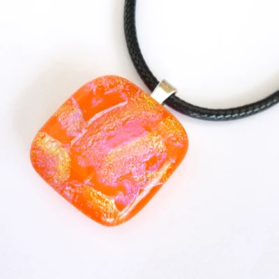 Handmade square orange dichroic glass pendant with 925 sterling silver bail and black cord necklace attached laying at a slight angle.