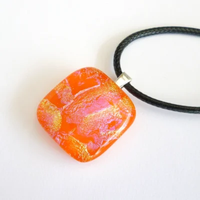 Orange dichroic glass pendant laying a an angle on a neutral background. 925 sterling silver bail and black cord necklace attached.
