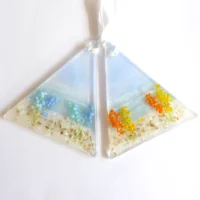 Two triangle shaped fused glass coastal suncatchers with flowers on. One with blue flowers and the other with orange & yellow flowers. Hanging from white ribbons.
