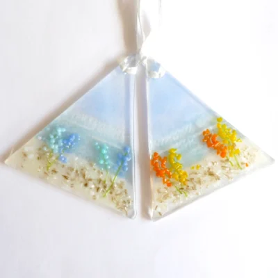 Two triangle shaped fused glass coastal suncatchers with flowers on. One with blue flowers and the other with orange & yellow flowers. Hanging from white ribbons.