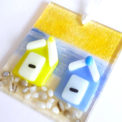 Yellow and blue beach huts fused onto glass. and hanging from a white ribbon. Suncatcher at an angle showing detail of glass.
