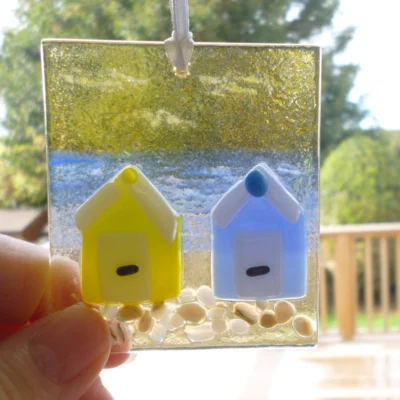 Square glass coastal suncatcher with yellow and blue beach huts fused on to glass. Yellow and blue background with frit for stones at the bottom.