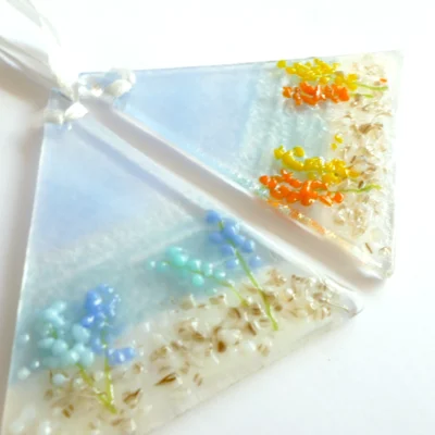 Two triangle shaped fused glass coastal suncatchers laying at a slight angle. One suncatcher has blue flowers and the other orange & yellow flowers. Hanging from white ribbons.