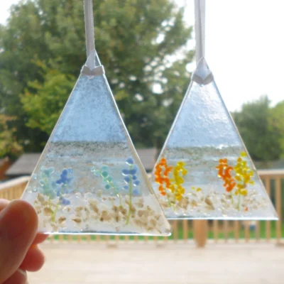 Two fused glass coastal suncatchers hanging in a window. Suncatchers have blue background, with a stones at the bottom and flowers.