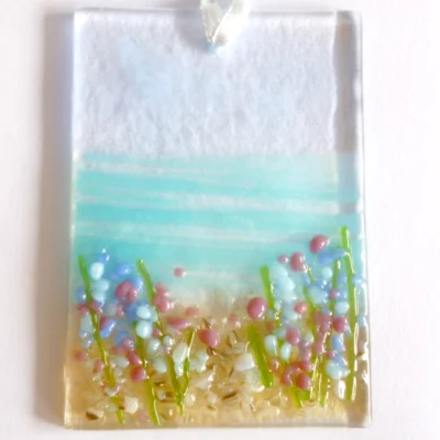 Small rectangle fused glass coastal suncatcher with blue background and blue & red flowers laying on a white background.