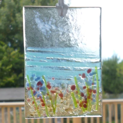 Small rectangle fused glass coastal suncatcher with blue and red flowers hanging from a white ribbon in a window.