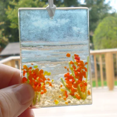 Small rectangle fused glass coastal suncatcher, being held at the corner, with flowers hanging from a white ribbon in a window.