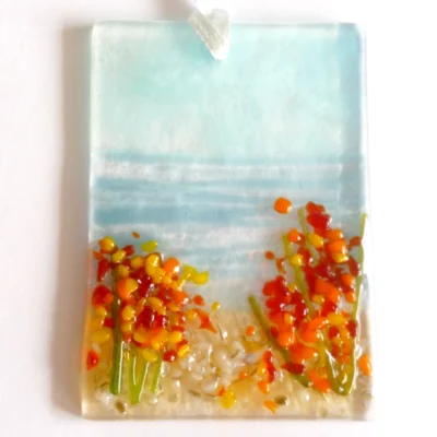 Small rectangle fused glass coastal suncatcher with blue background and yellow and orange flowers laying on a white background.