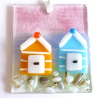 Front view of fused glass coastal suncatcher. Orange and blue stripe beach huts fused onto a layer of glass with pin and blue background.