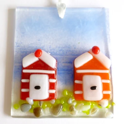 small square fused glass coastal suncatcher with two beach huts in red and orange with white stripes. Blue background for the sky and frit for sand and grass