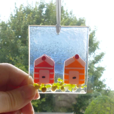 Fused glass coastal suncatcher with two beach huts in orange and red being held at the corner.