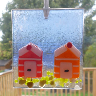 Square fused glass coastal suncatcher hanging in a window with light coming through it. Red and orange beach huts with white stripes