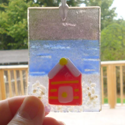 Small fused glass suncatcher being held by the bottom left corner. Small red and yellow beach hut fused onto a rectangle shaped piece of glass.