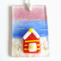 Small fused glass suncatcher with red and yellow beach hut hanging from a white ribbon. Background of suncatcher is pink blue and white.
