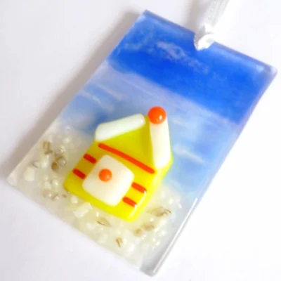Small fused glass coastal suncatcher laying at an angle on a white background. Yellow beach hut with red stripes and blue sky & sea.