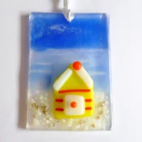 Small fused glass suncatcher with a yellow beach, hanging from a white ribbon. Beach hut has red stripes and white roof.