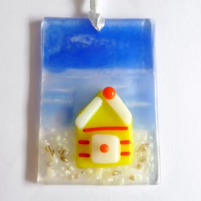 Small fused glass suncatcher with a yellow beach, hanging from a white ribbon. Beach hut has red stripes and white roof.