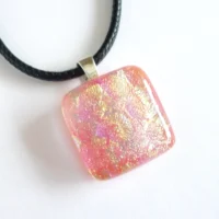 Square dichroic glass pendant laying at a slight angle on a white background. Dichroic has shimmers of pink and yellow / gold on top. Sterling silver bail and black cord necklace attached.