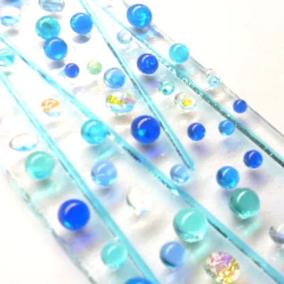 Close up of blue fused glass suncatcher with blue / turquoise beads and stringers and dichroic beads. Tactile design hanging from a clear cord.
