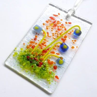 Blue glass flower suncatcher laying at a slight angle. Quirky square light and dark blue flowers, with green stems and orange and green frit for decoration.