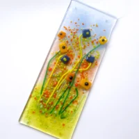 Blue and orange fused glass flower suncatcher laying at an angle. Background of light blue, orange and green with quirky square orange and blue glass flower with long stems.