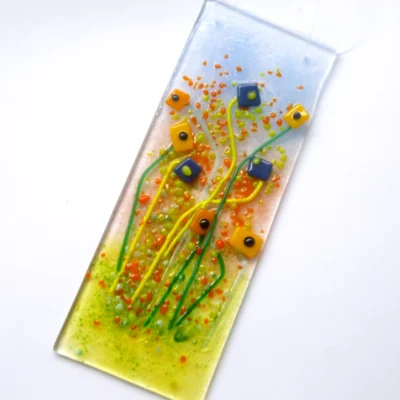 Blue and orange fused glass flower suncatcher laying at an angle. Background of light blue, orange and green with quirky square orange and blue glass flower with long stems.