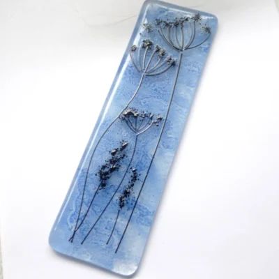 Blue fused glass dandelion suncatcher laying at an angle on a neutral background.