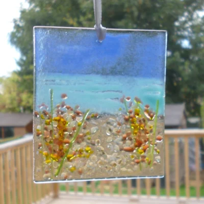 Rectangle handmade fused glass coastal suncatcher with deep red and white flowers on a beach. Hanging from a white ribbon in a window with natural light coming through.