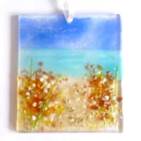 Rectangle fused glass coastal suncatcher laying on a white background. Blue sky, turquoise sea and red & white flowers on the beach. Hanging from a white ribbon.