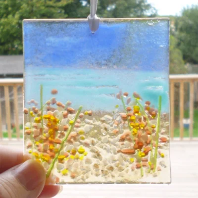 Fused glass coastal suncatcher hanging in a window with natural light coming through, being held at the bottom left corner.