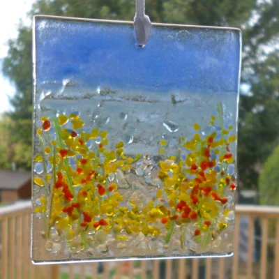 Rectangle fused glass coastal suncatcher with red, yellow and blue flowers hanging in a window with natural light coming through. Hangs from a white ribbon.